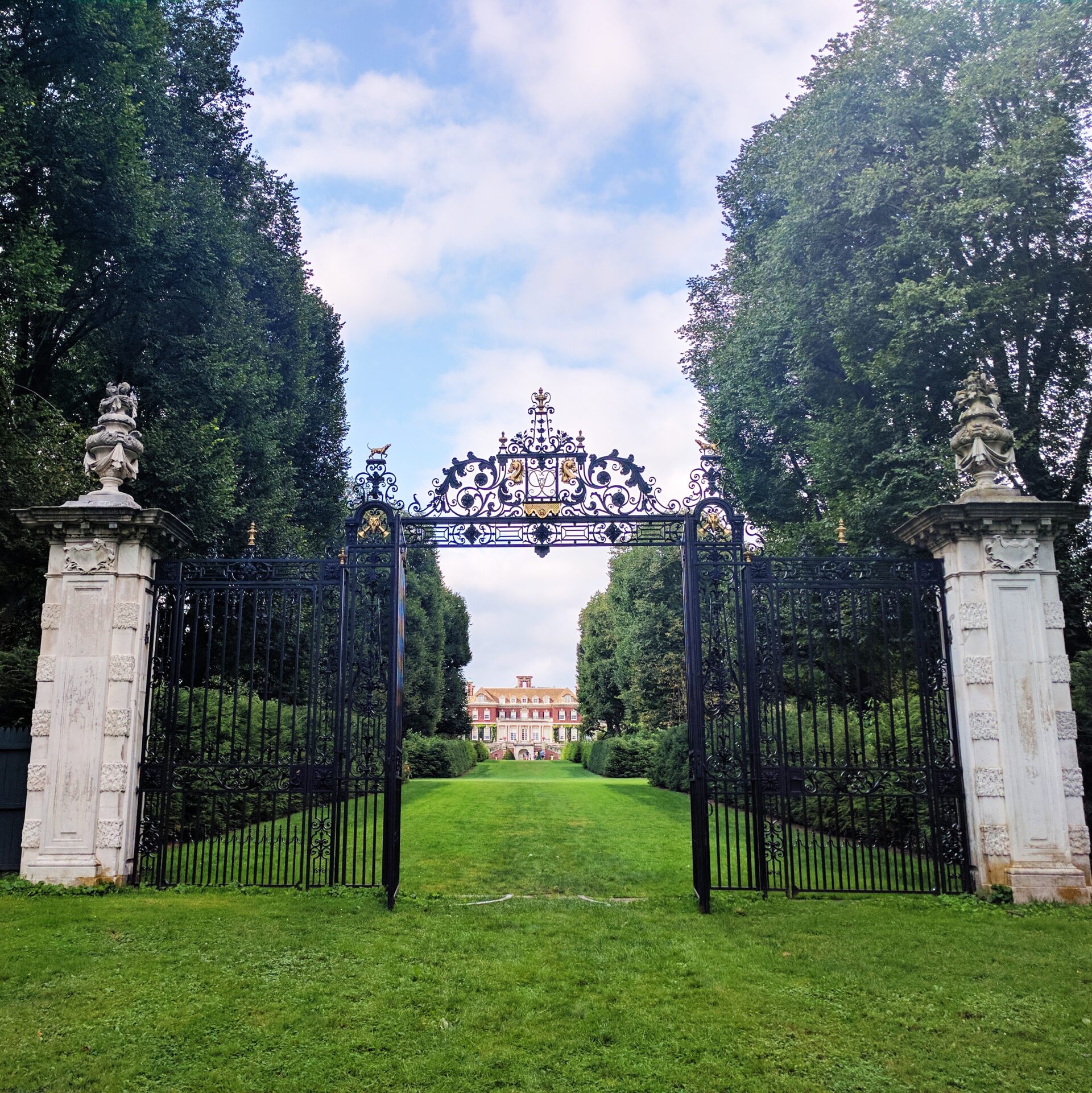 an intricate gate standing in the grass with a grand house visible on the other end